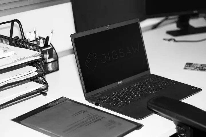 Desk at the Jigsaw office with a laptop showing the Jigsaw logo, document trays and paperwork