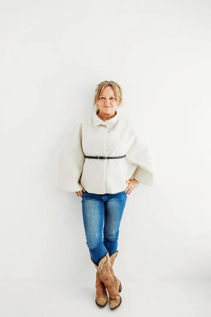 Emma Kneale standing against a white wall in a cream jumper, jeans and cowboy boots